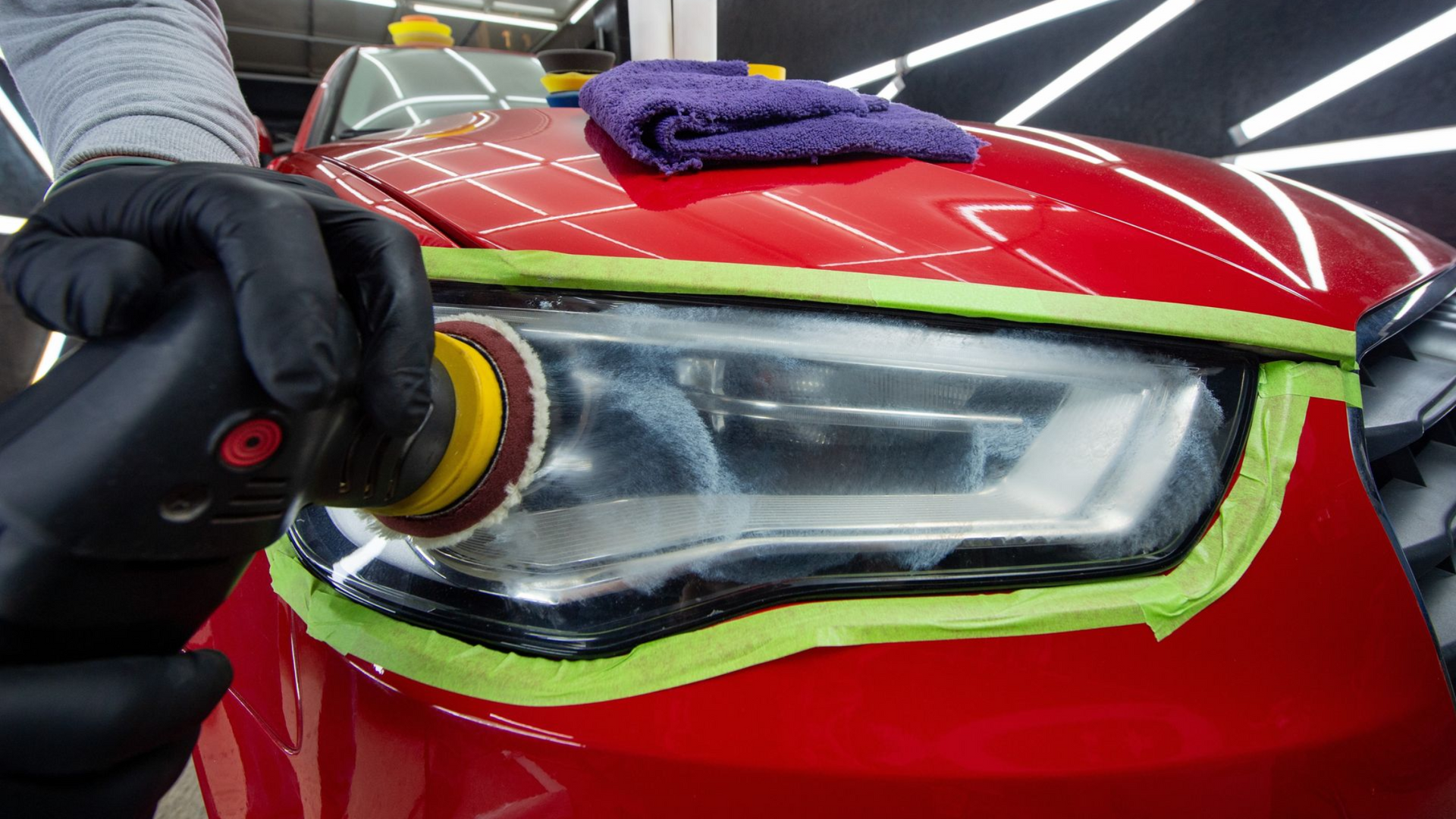 Gloved hand polishing a red car’s headlight with a yellow cloth