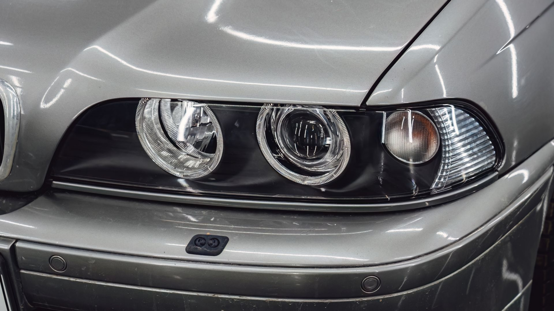 Close-up of a silver car’s front headlight assembly with three circular lamps and a black grille.