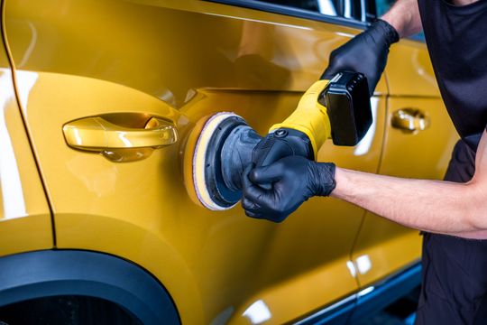 Gloved worker polishing a yellow car door with a buffer in an auto shop
