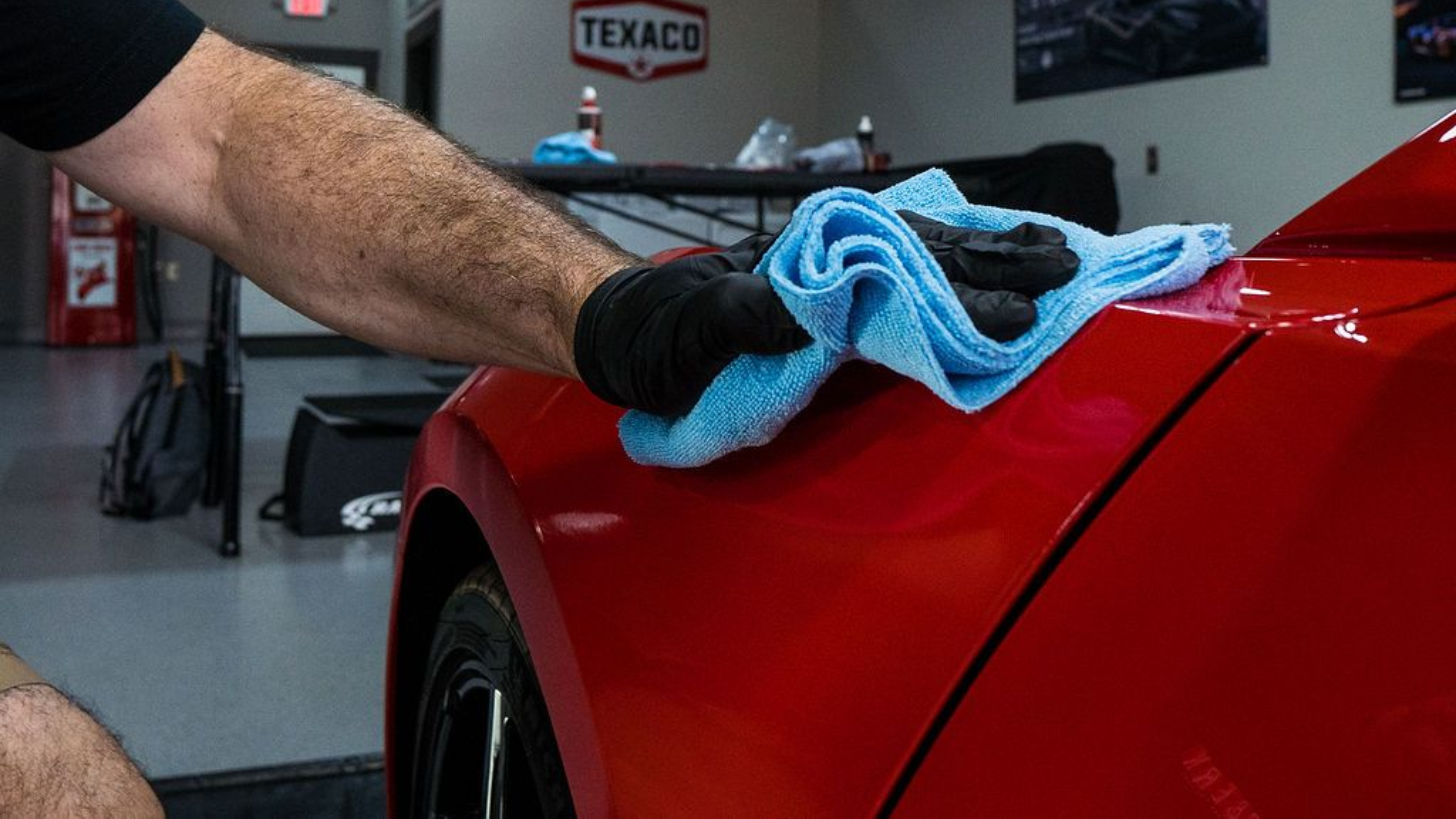 Person wiping a red car hood with a blue microfiber cloth in a garage