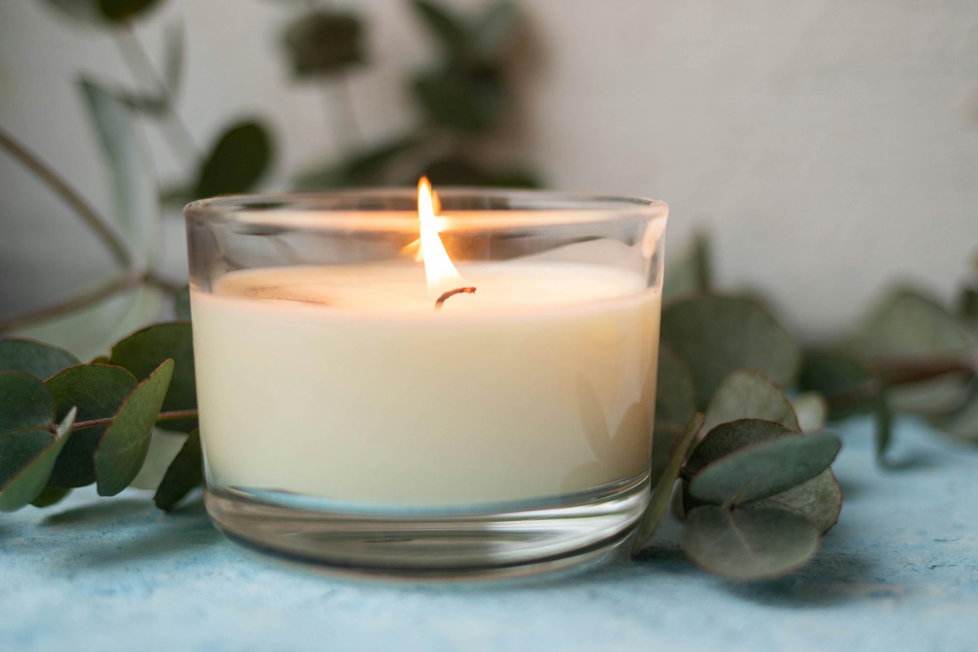 White candle in a glass jar with a small flame, surrounded by green leaves on a soft blue surface.
