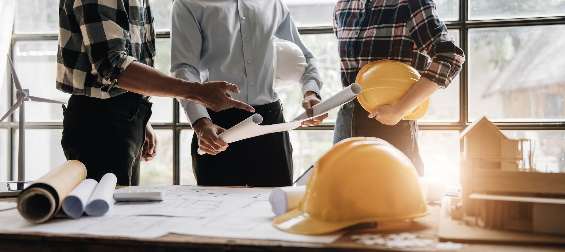 Trois personnes, probablement des ingénieurs, examinant des plans à une table, portant des casques de chantier jaunes, à l'intérieur, près d'une fenêtre.