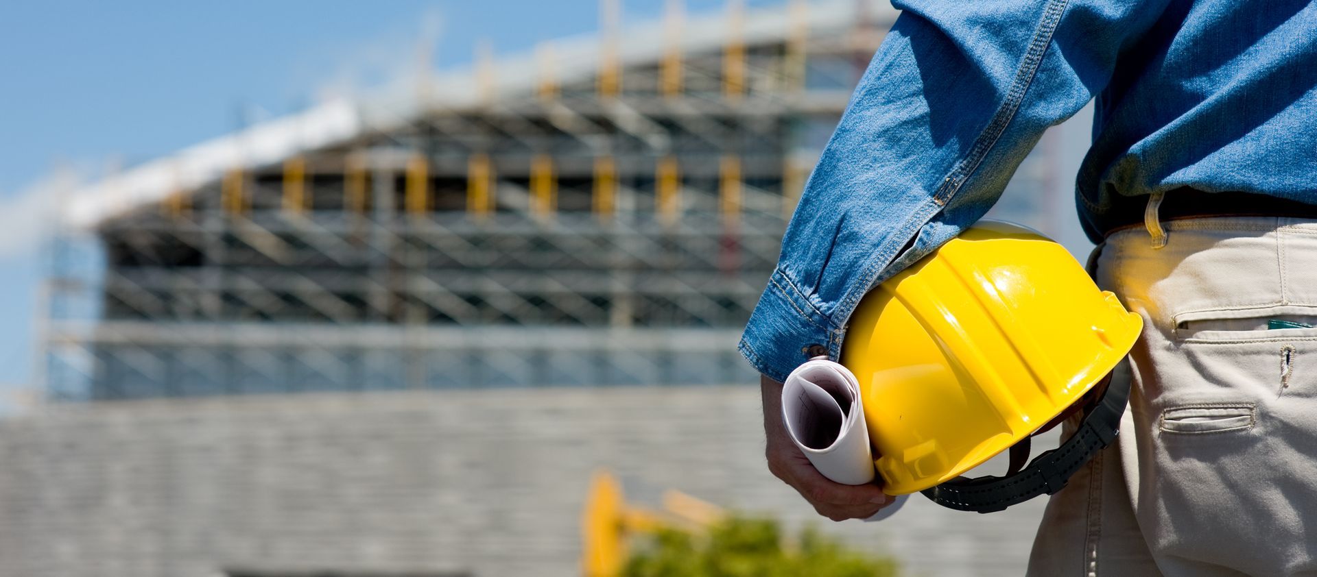 Ouvrier du bâtiment tenant un casque jaune et des plans roulés, avec un bâtiment en construction en arrière-plan.