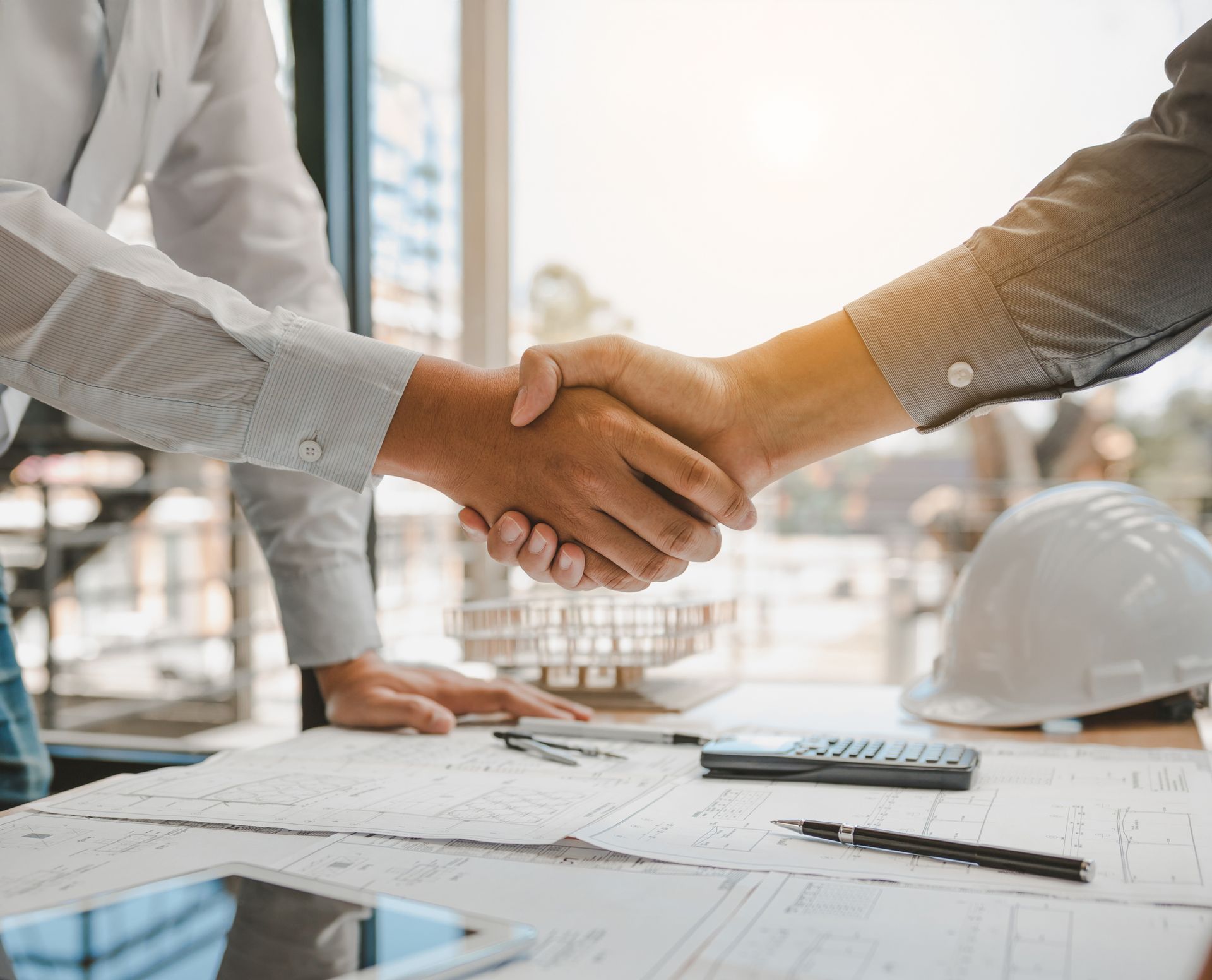 Two people shaking hands over blueprints and a calculator at a desk.