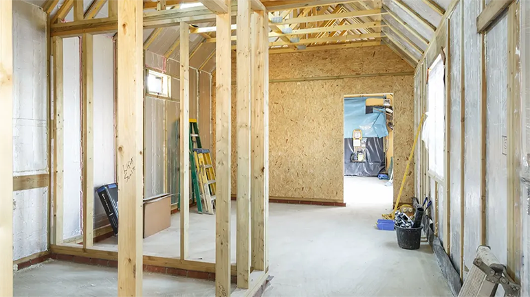 Intérieur d'un bâtiment en construction, avec charpente en bois visible, murs non finis et sol en béton.