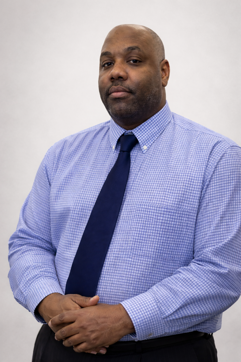 A professional portrait of a person in a blue-checkered button-down shirt and navy blue tie standing against a white wall.