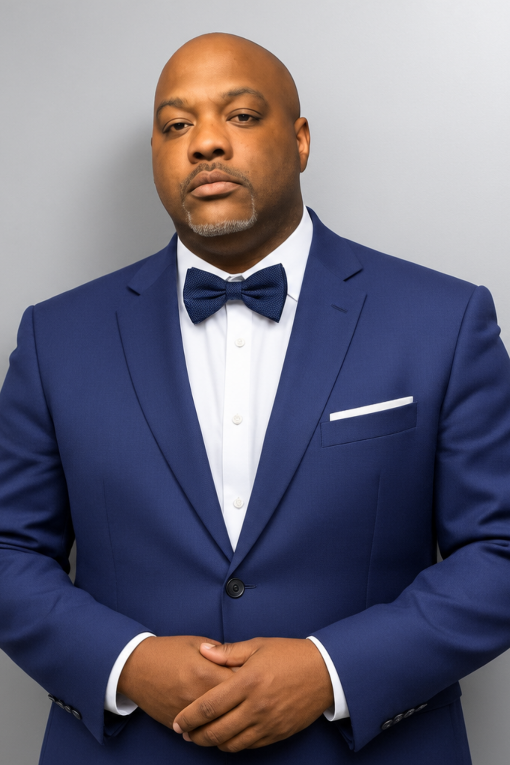 A professional portrait of a person in a blue-checkered button-down shirt and navy blue tie standing against a white wall.