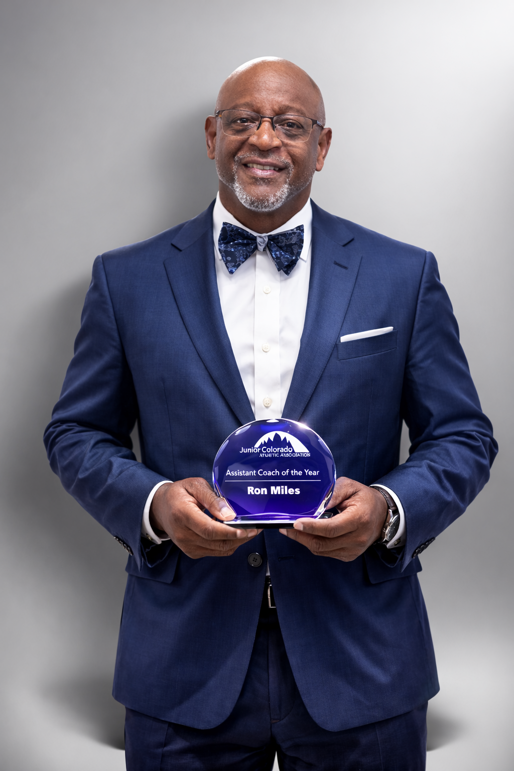A smiling person in a blue suit and patterned bow tie holds a round award plaque against a plain gray background.