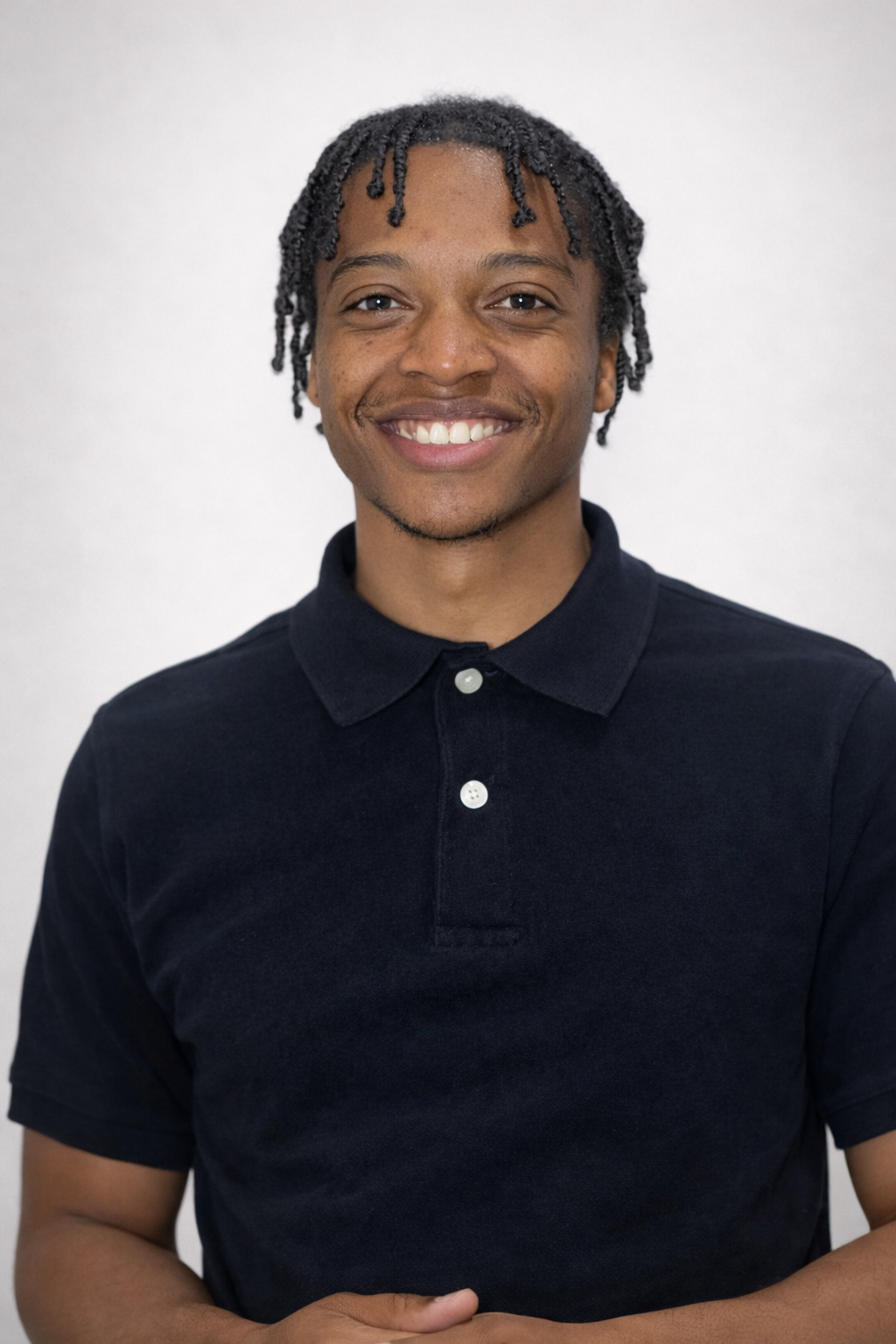 A person with braided hair and a friendly smile wears a dark blue polo shirt against a light grey background.
