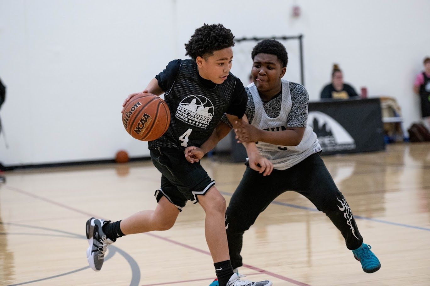 A basketball player in a black jersey dribbles the ball past a defender in a light gray jersey on an indoor court.