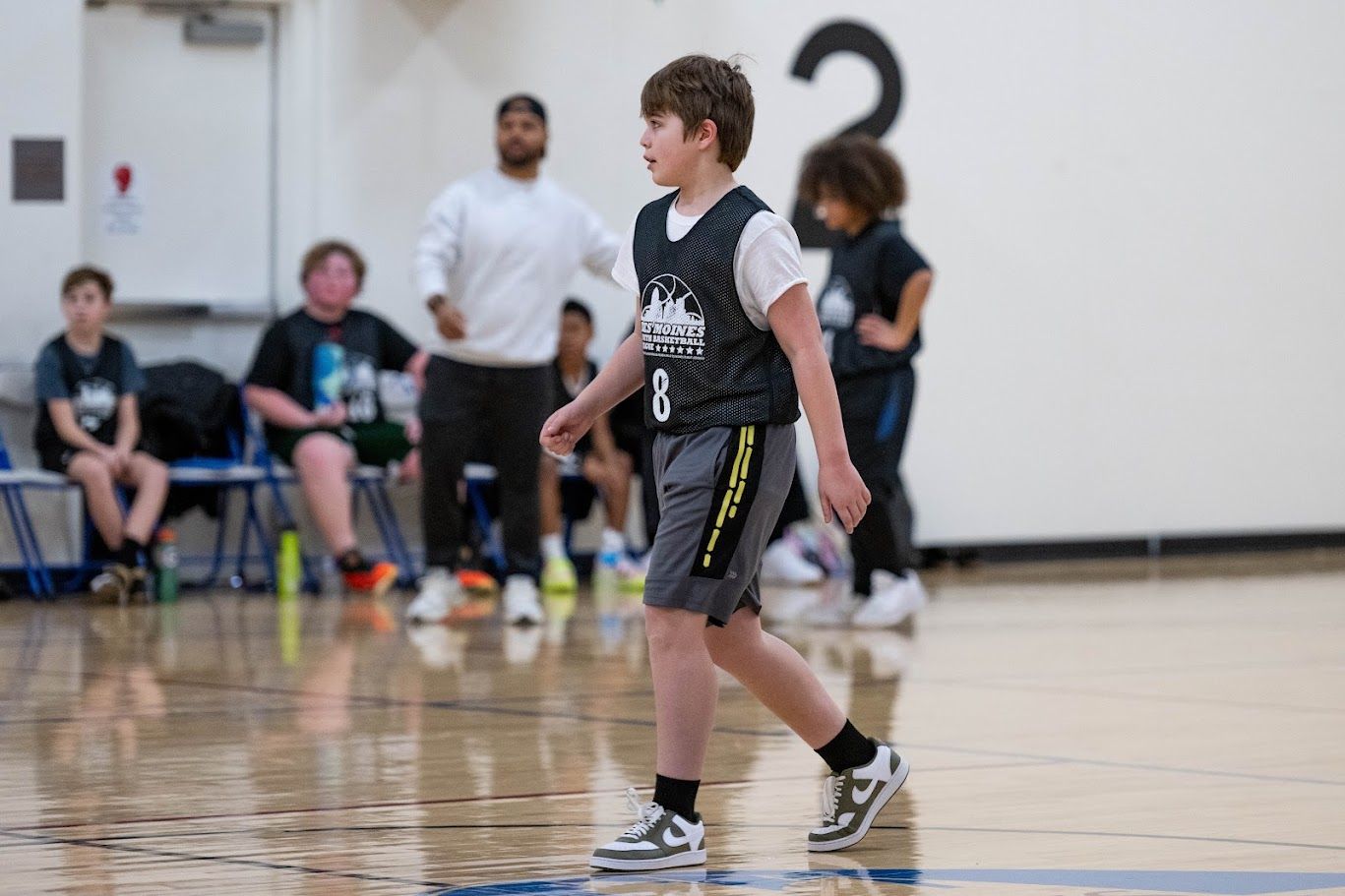 A person in a black jersey and shorts walks across a basketball court with other players and an adult in the background.
