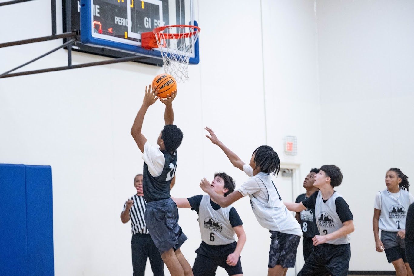 A basketball player in a dark uniform jumps to shoot, defended by two teammates in light jerseys during a game.