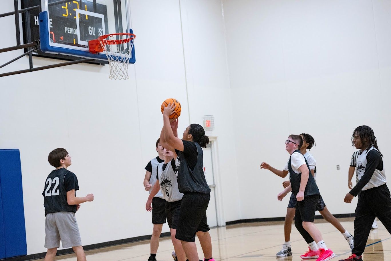 A group of youth basketball players in a gym, with one player jumping to shoot the ball toward the hoop.