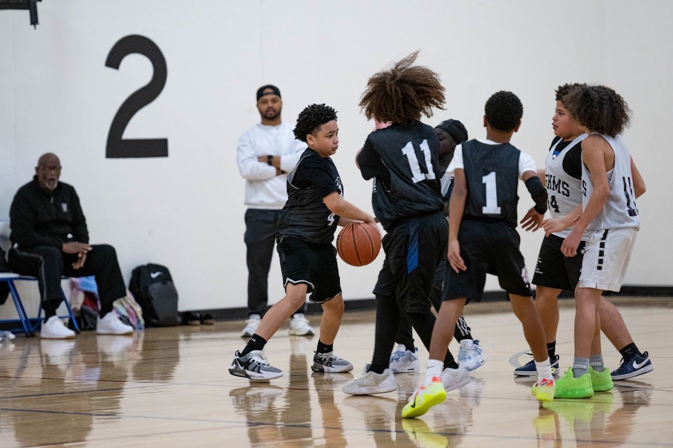 Young players in dark and white uniforms compete for a basketball on an indoor court while adults watch from the side.