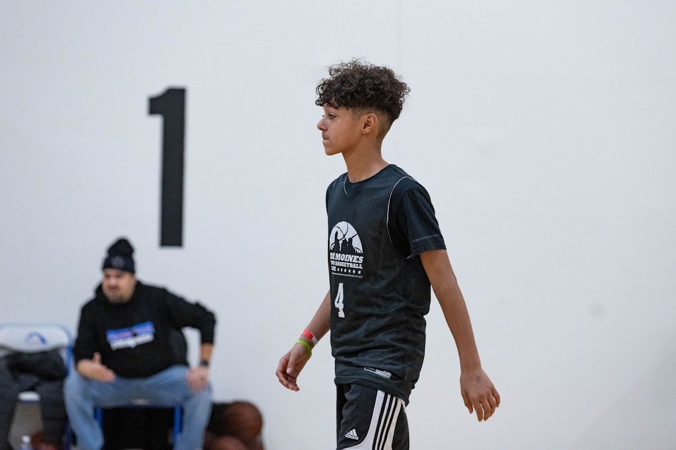 A player in a patterned black jersey walks across a basketball court in front of a white wall with a large black