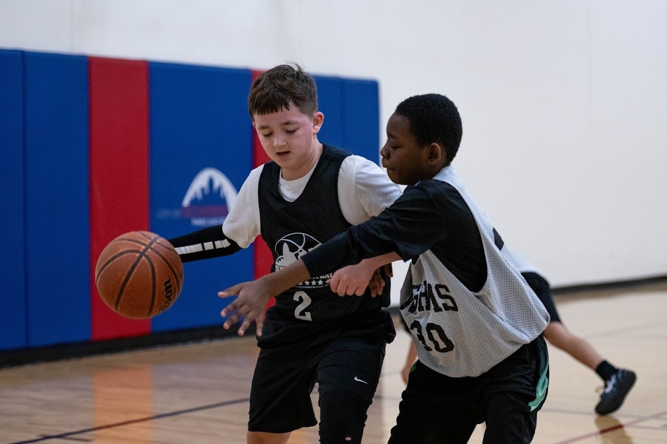 Two youth basketball players wearing numbered jerseys compete for the ball on an indoor court with blue wall padding.