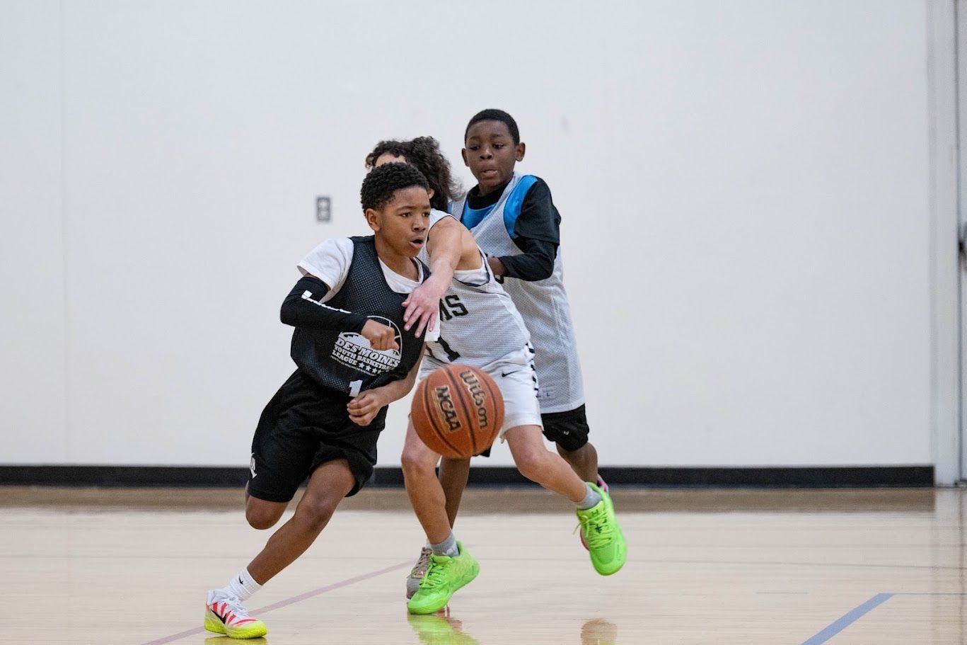 Three youth basketball players in jerseys compete on a court, with one dribbling the ball while the others defend.