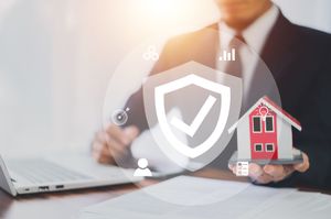 Businessman holding a small house model beside a laptop, with a shield security icon overlay.