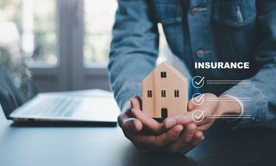 Hands holding a small house model beside a laptop, with “INSURANCE” text on screen.
