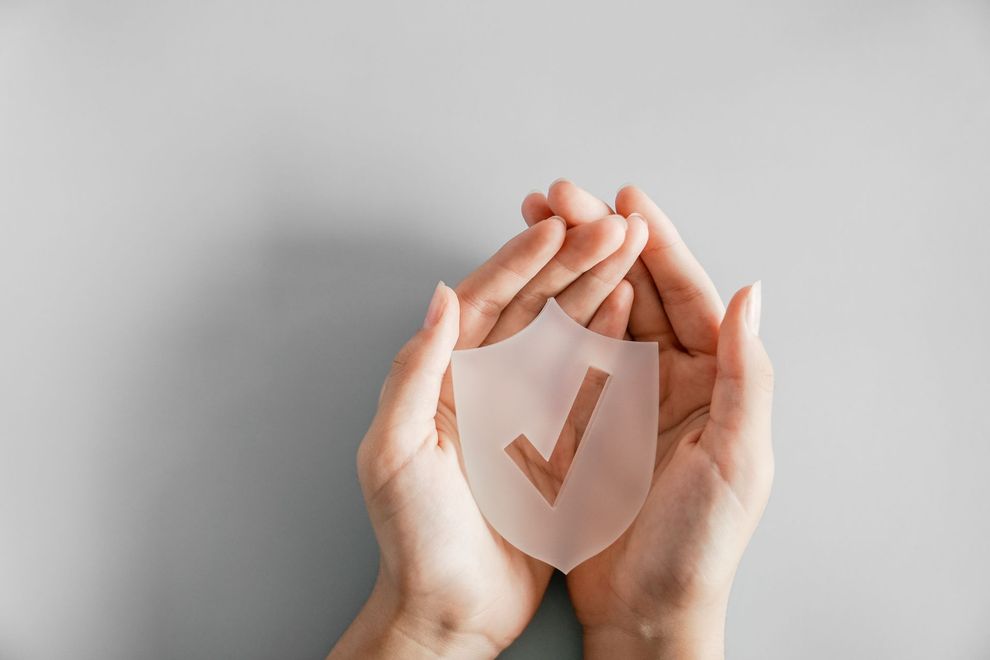 Hands cupping a translucent shield with a check mark on a gray background