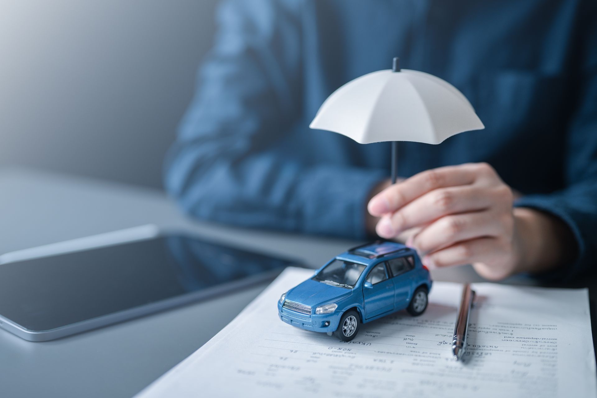 Hands holding a white umbrella over a blue toy car on documents beside a tablet and pen