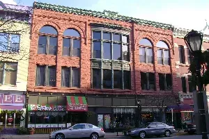 Brick storefront building with arched windows and striped awnings on a city street, cars parked below