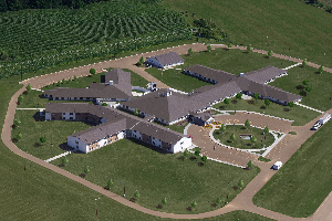 Aerial view of a large farmhouse-style building with a circular driveway, green lawns, and surrounding farmland.