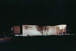 Nighttime view of a modern white building lit from below, with a small tree in front and dark surroundings