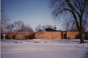Snow-covered park with a brick building and bare trees under a clear blue sky