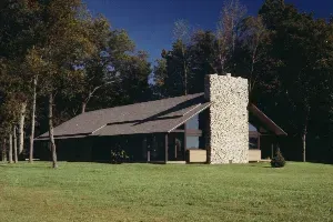 Small dark cabin with a stone chimney on a grassy lawn, surrounded by trees.