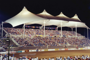 Crowded nighttime rodeo arena under white canopy tents with bright lights and grandstands