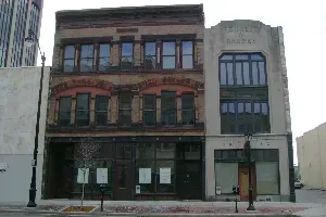 Old brick storefronts on a city street, with a faded gray building and snow along the sidewalk.