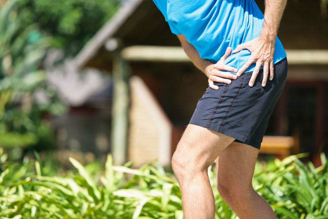 Person in blue shirt and black shorts holding their lower back outdoors in a garden.