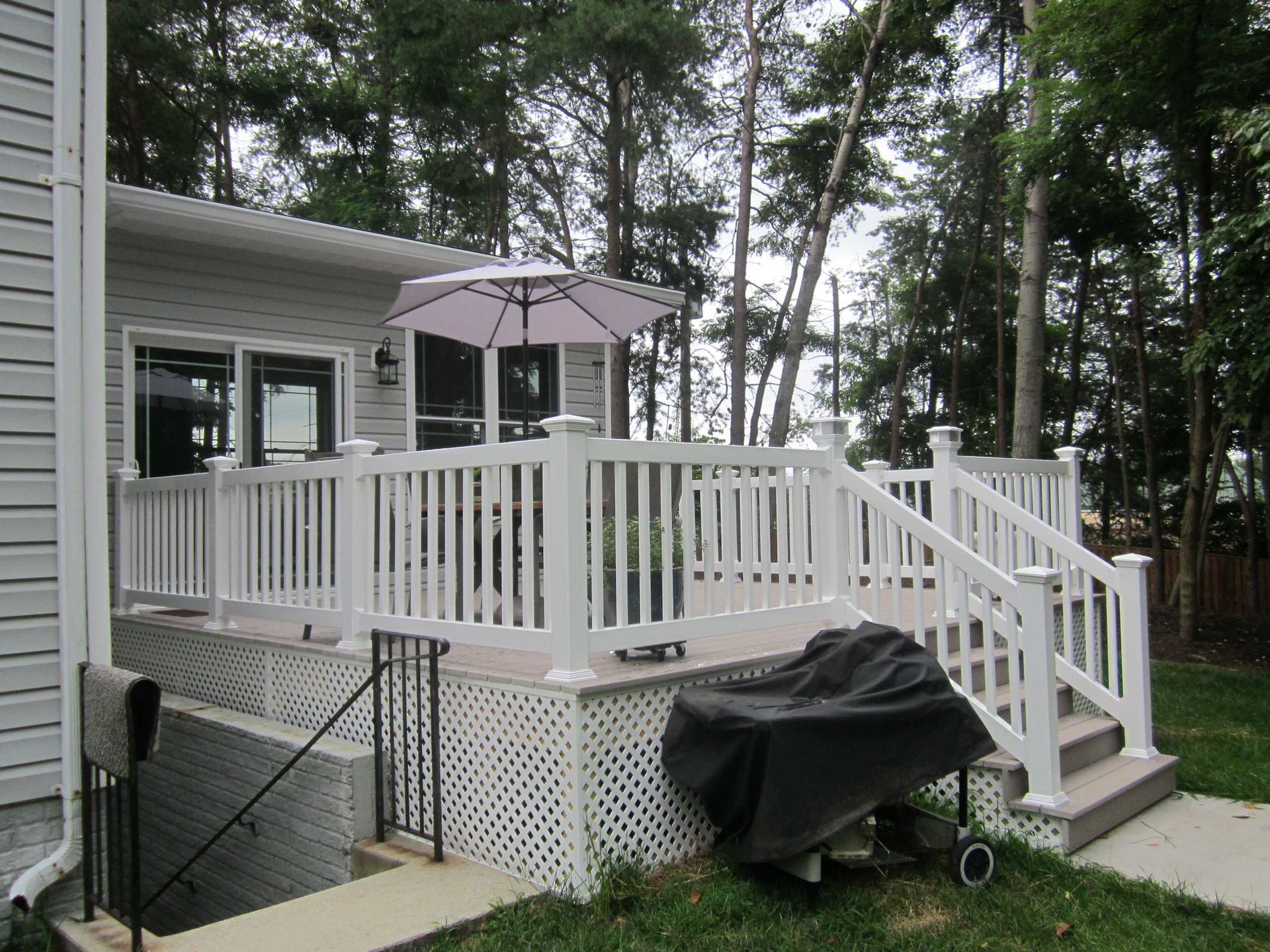 Back deck with white railing, gray house, patio umbrella, and covered grill in a wooded yard