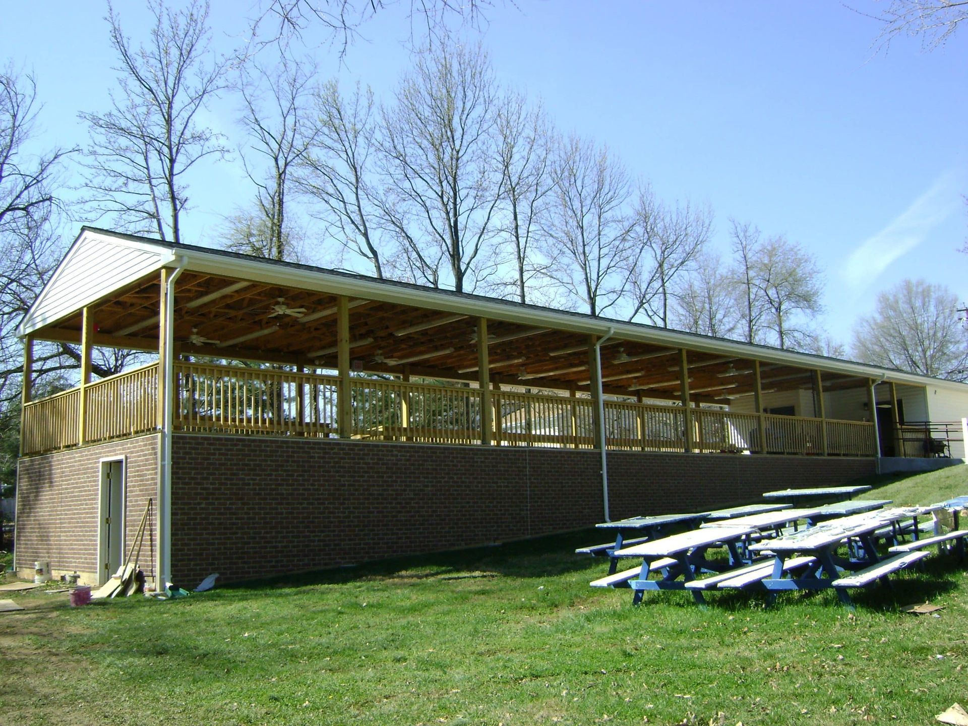 Covered outdoor pavilion beside grassy field with bleachers and leafless trees under a blue sky