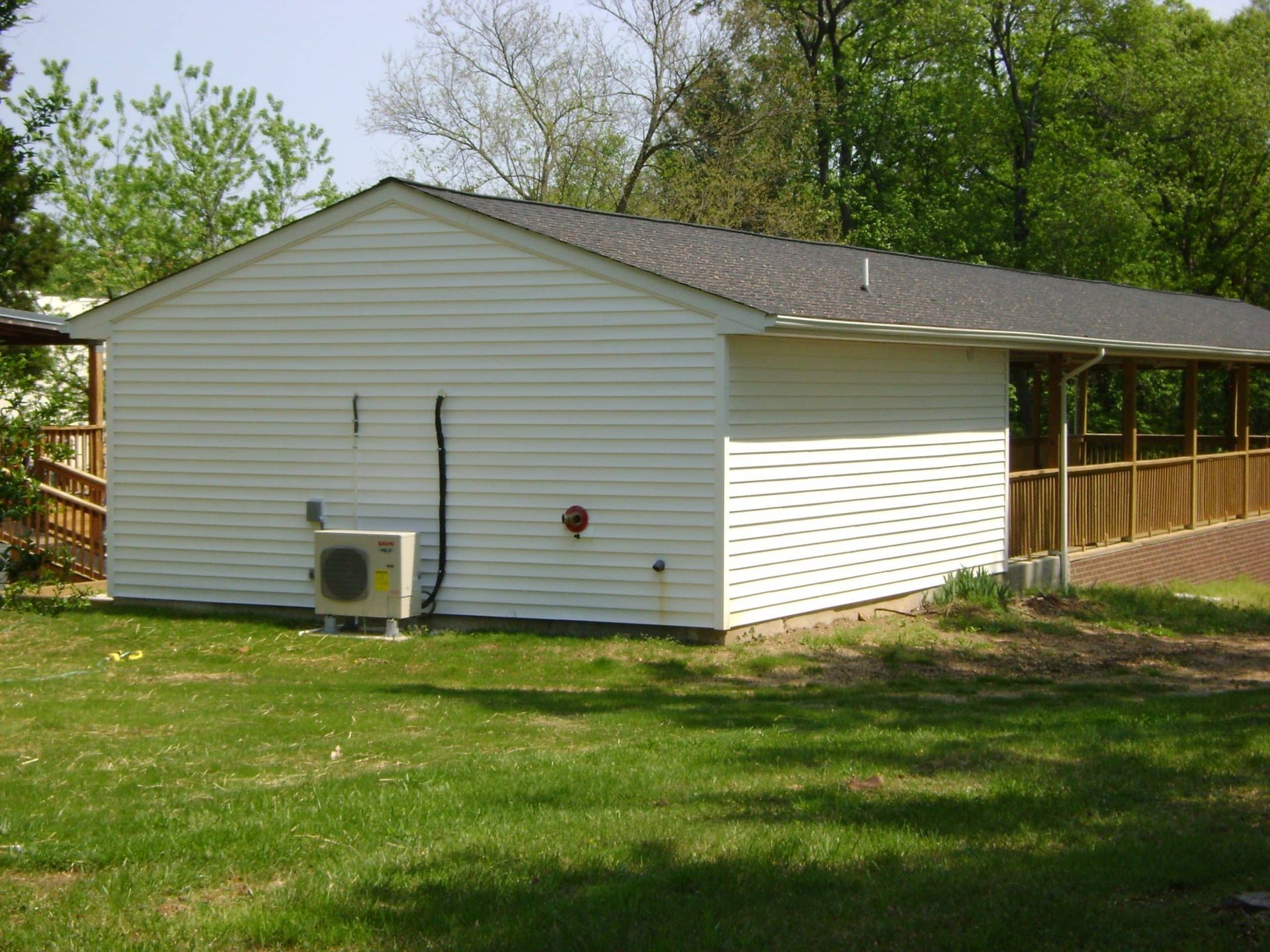 White house with gray roof and attached wooden deck in a grassy yard, trees in the background