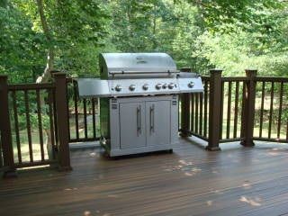 Stainless steel gas grill on a wooden deck surrounded by green trees and railings