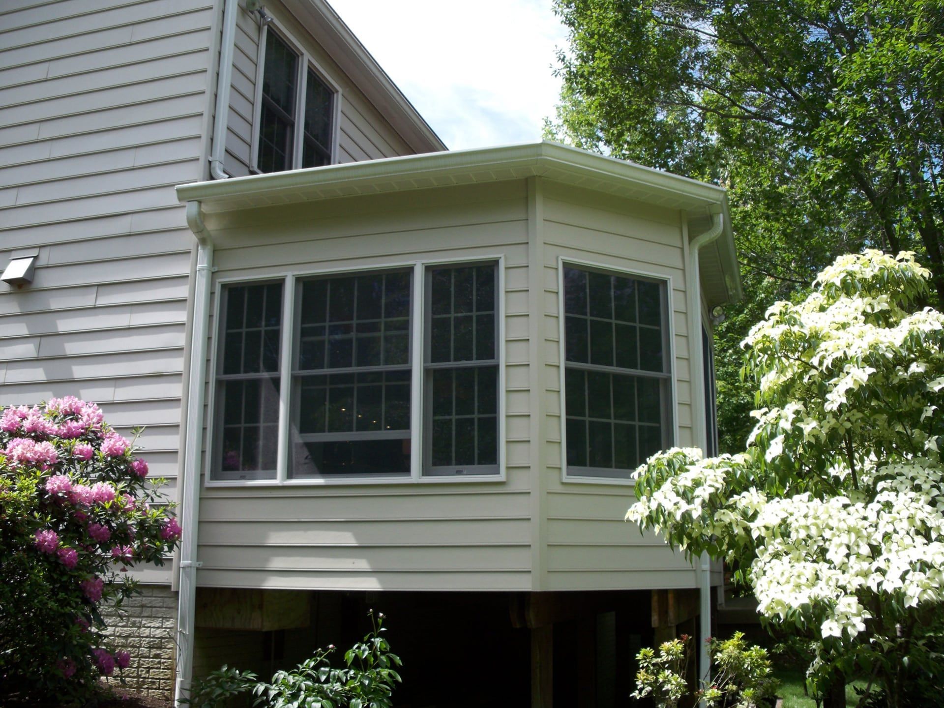 White enclosed porch with windows attached to a gray house, surrounded by flowering shrubs and trees