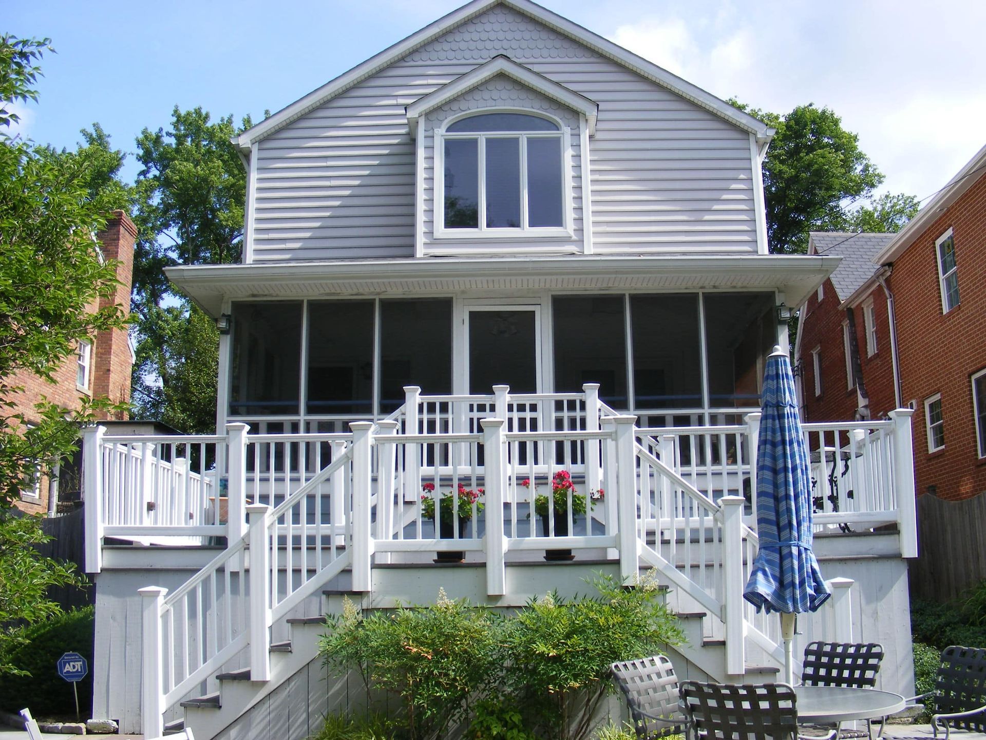 White two-story house with a front porch, white railings, and stairs, flanked by brick buildings.