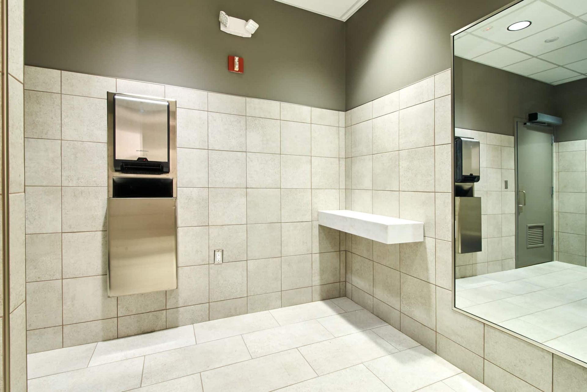 Modern restroom with white tile walls, a stainless steel dispenser, sink shelf, and large wall mirror