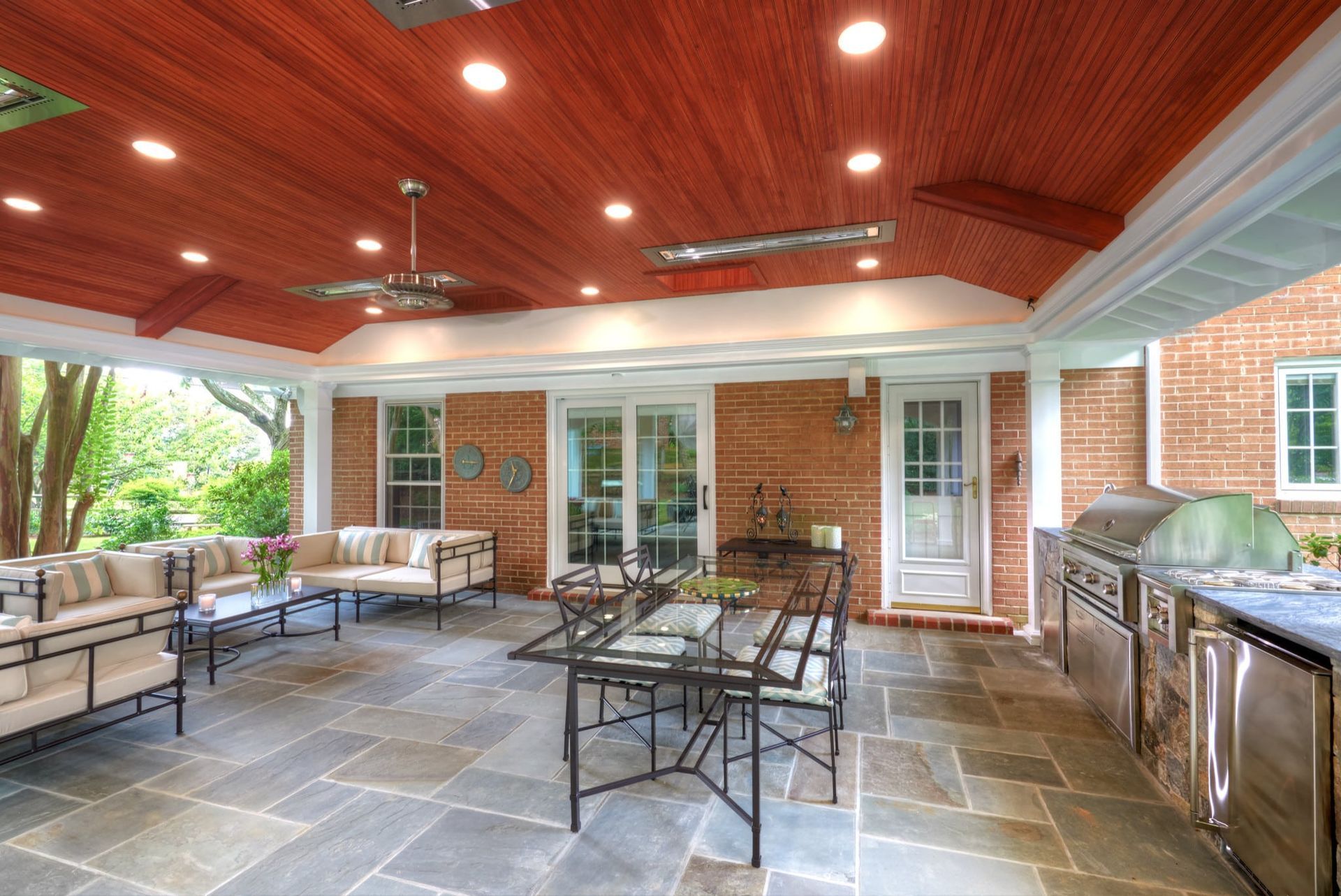 Covered patio with stone floor, red wood ceiling, white beams, seating, and a grill beside brick walls.