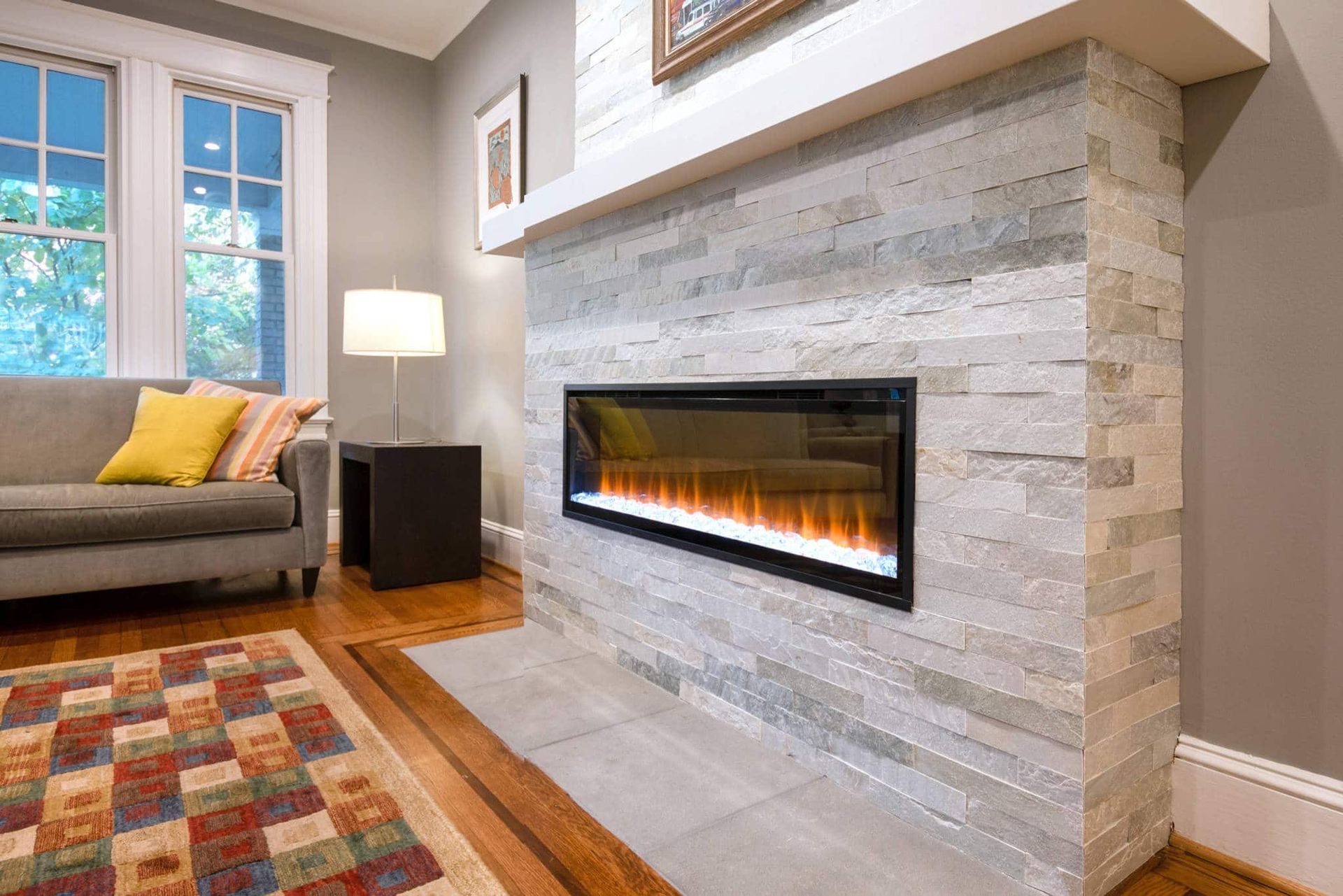 Modern living room with a stone fireplace, lit fire, sofa, lamp, and large windows