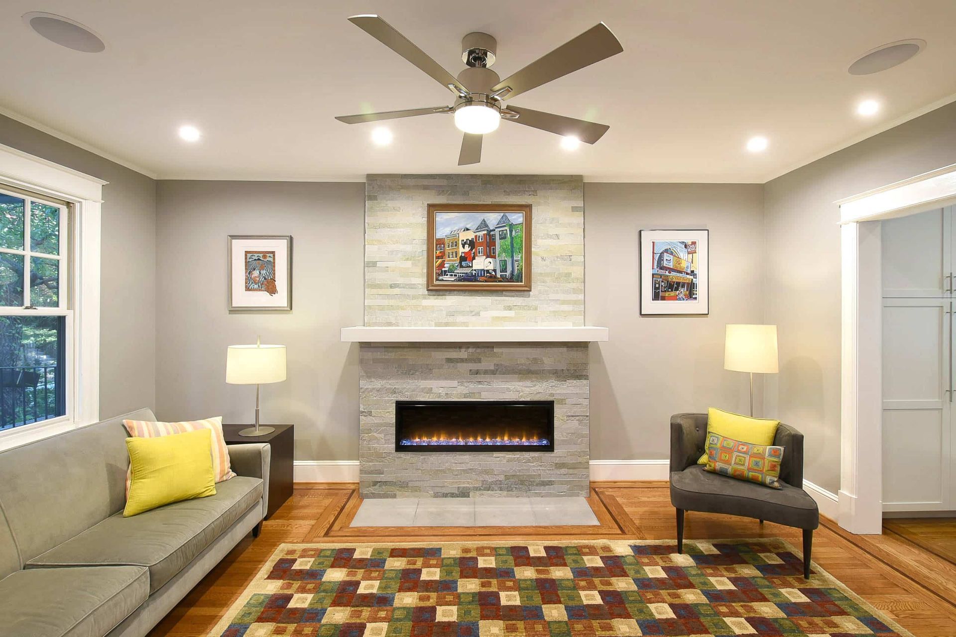 Bright living room with fireplace, sofa, armchair, colorful rug, lamps, and ceiling fan.