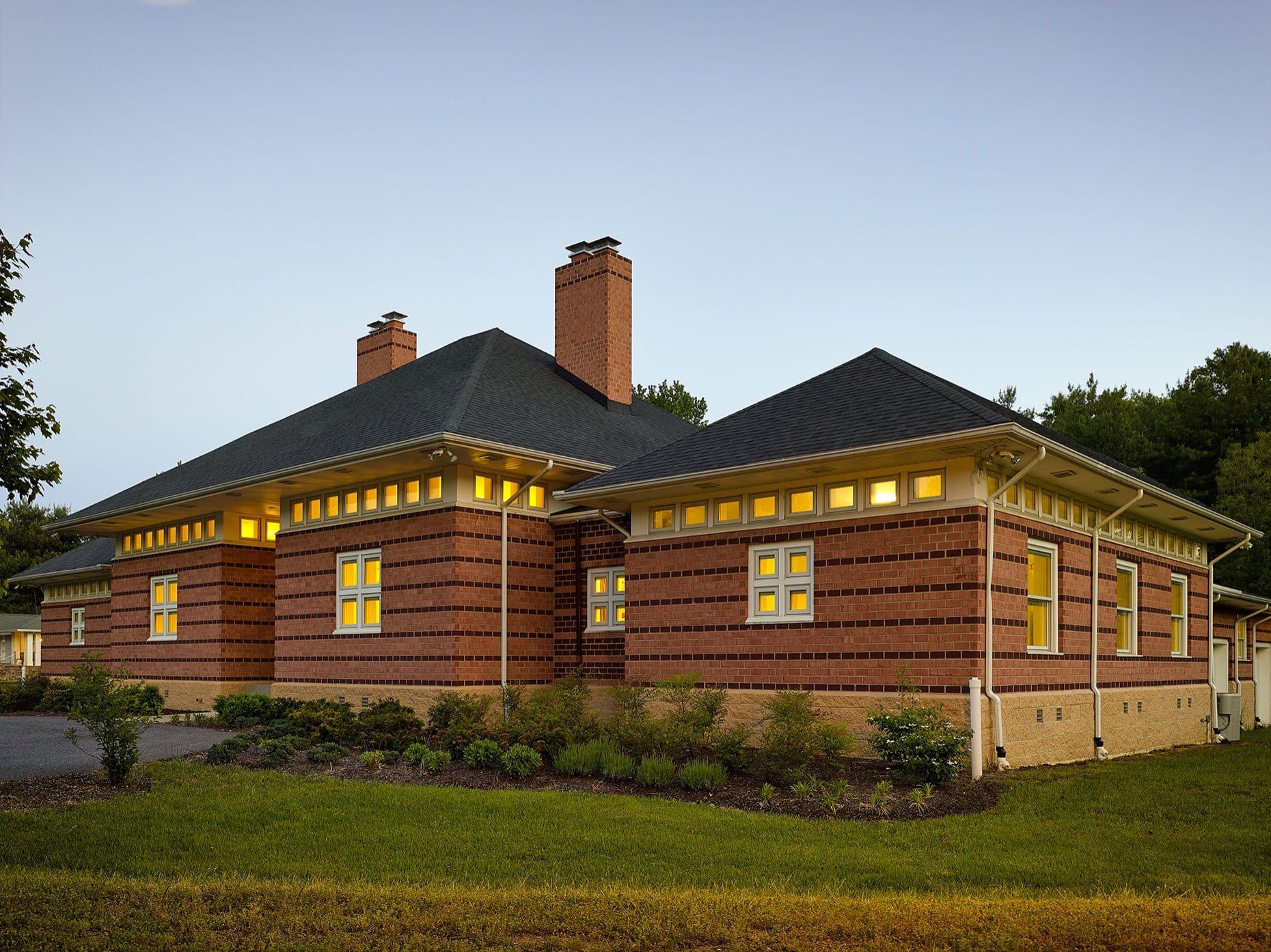 Single-story brick house with lit windows and dark roof at dusk, surrounded by grass and trees
