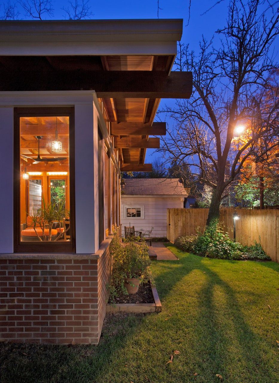 Backyard at dusk with a lit house window, green lawn, fence, trees, and a streetlight glowing nearby