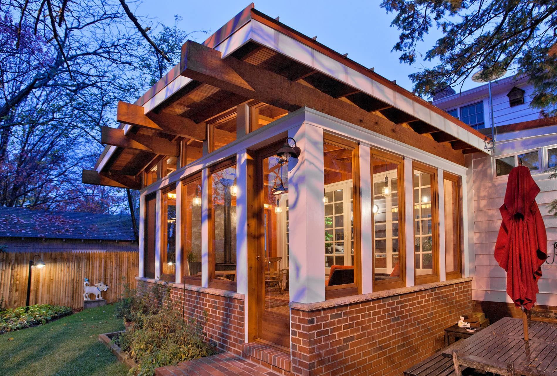 Brick sunroom with large windows at dusk, lit warmly, beside a fenced yard and red patio umbrella