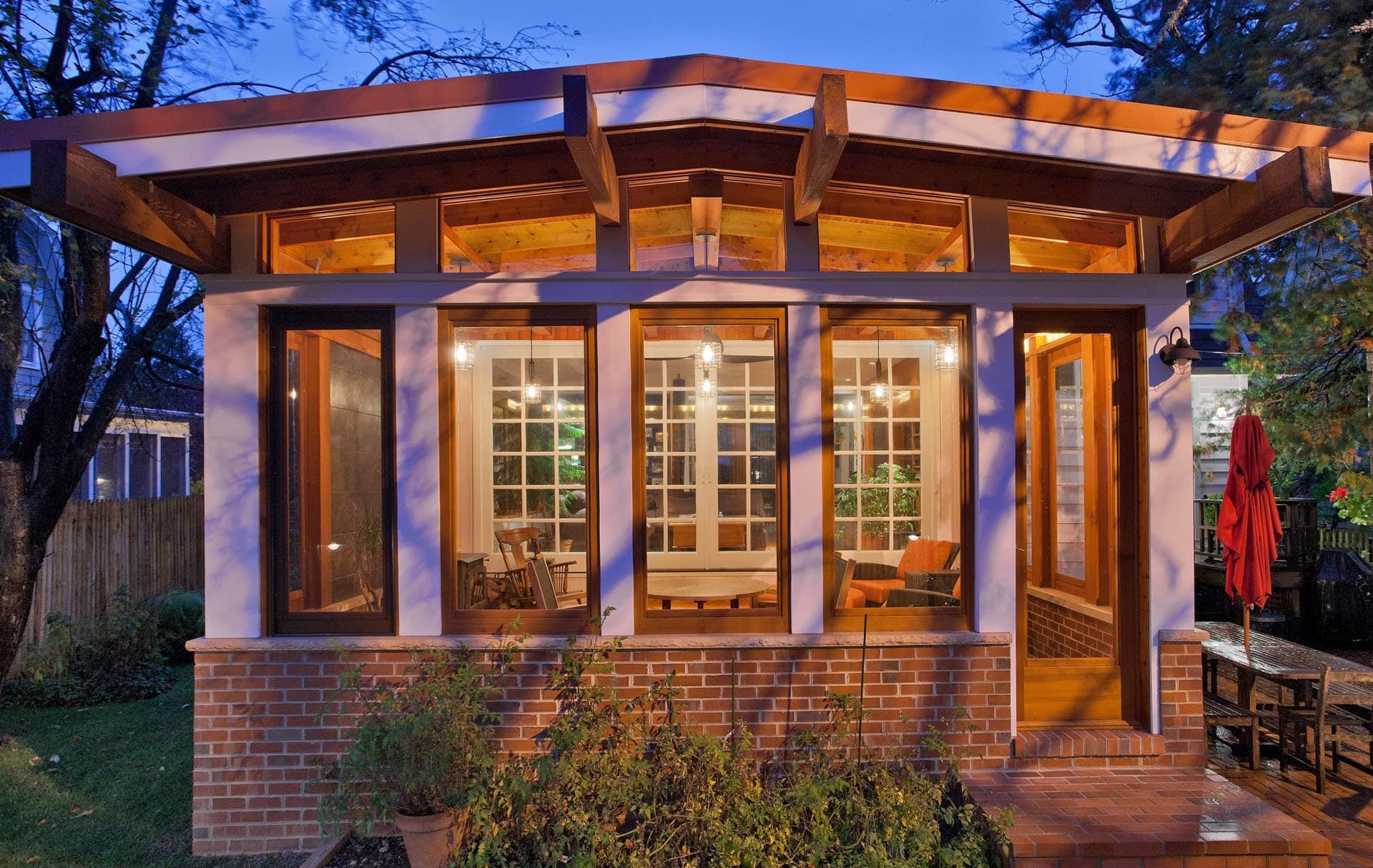 Wooden pavilion with large windows and brick base, lit warmly at dusk in a garden courtyard