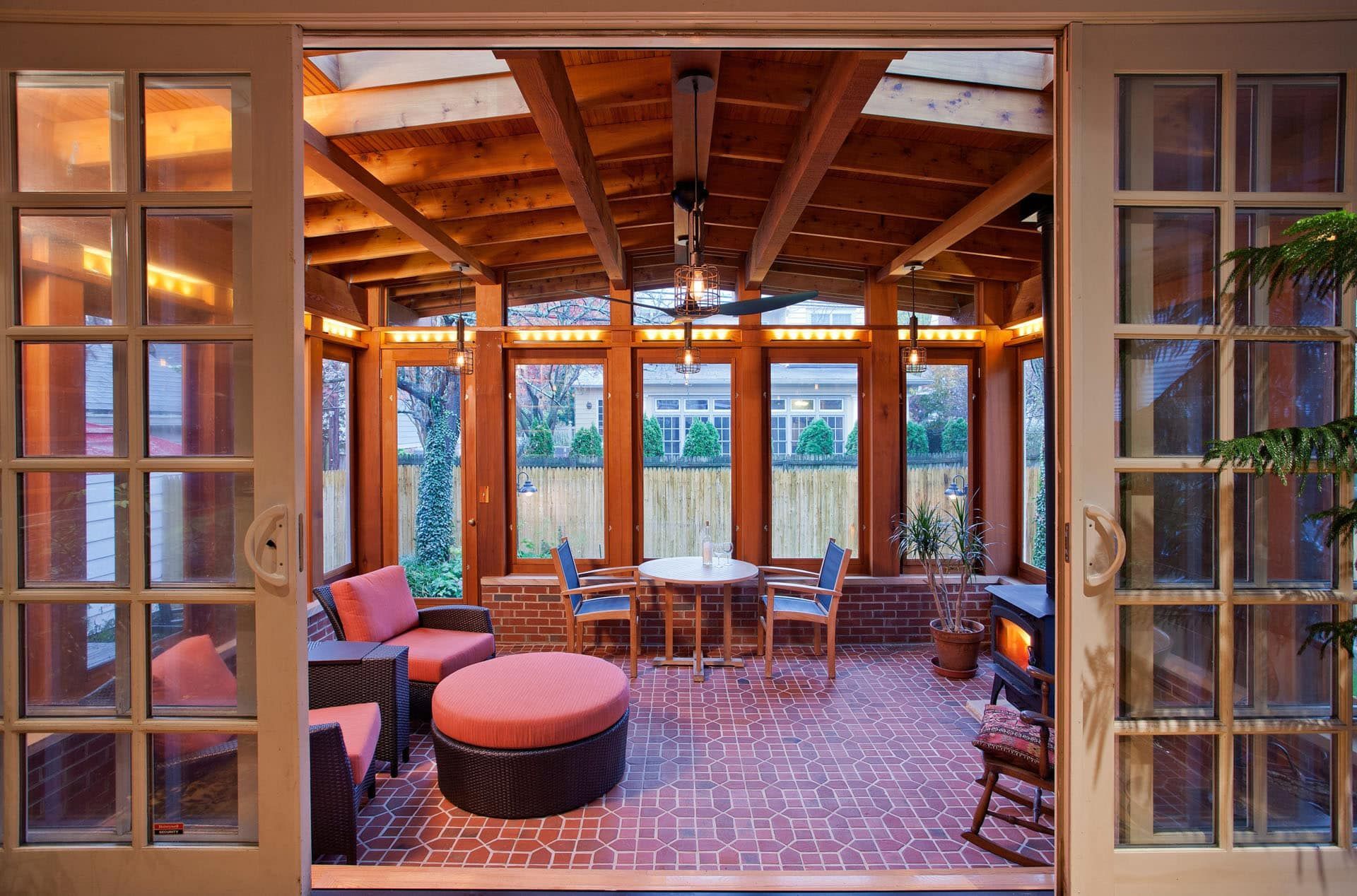 Open-air patio lounge with brick floor, wooden beams, cushioned seating, and glass doors at sunset