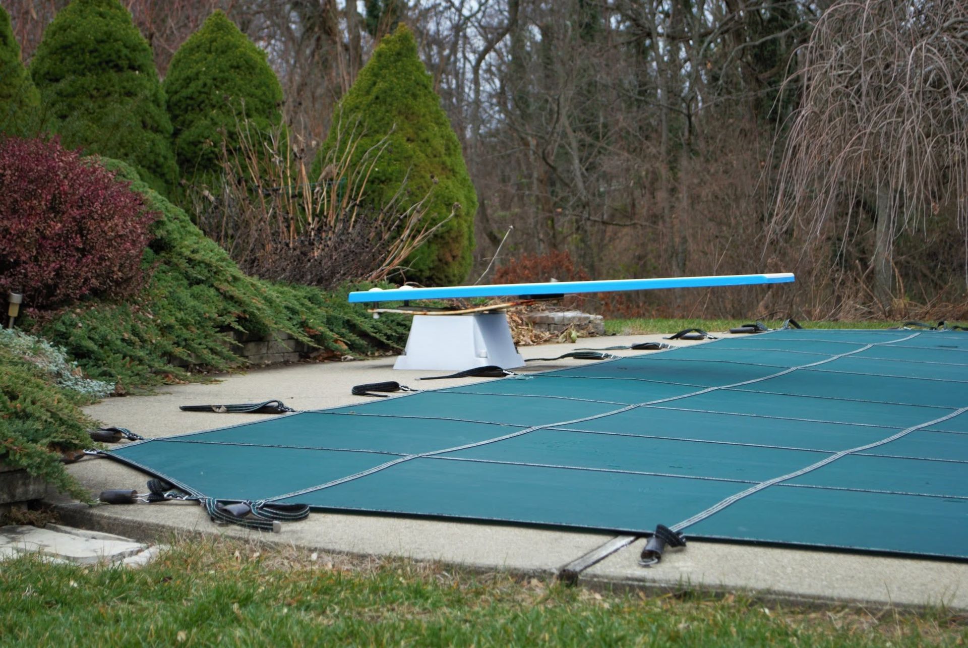 Empty outdoor swimming pool with a blue cover and diving board surrounded by trees