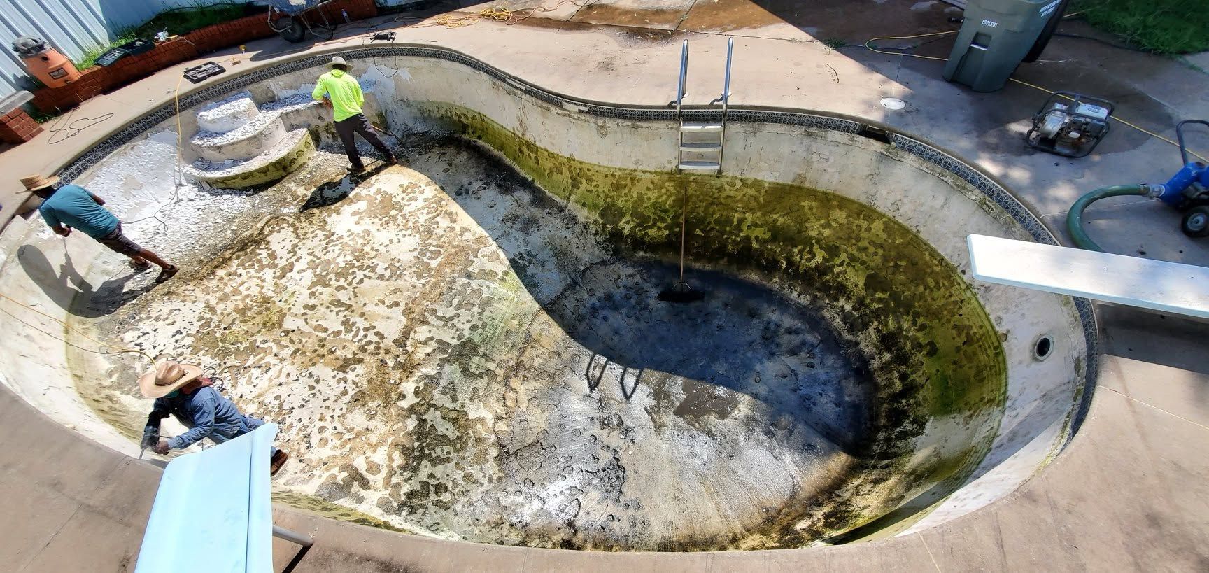 Workers cleaning a drained, algae-lined swimming pool under repair